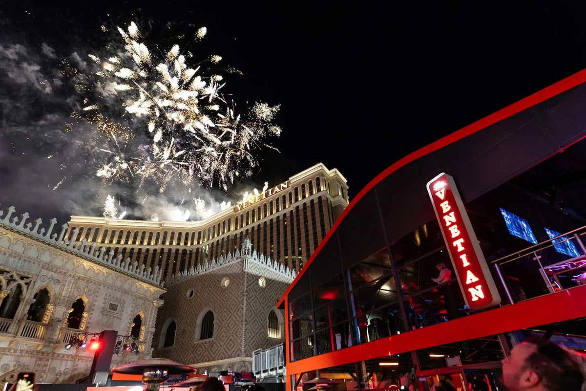 Nighttime scene on the Las Vegas Strip with fireworks in the sky above The Venetian hotel, viewed from street level with a red, glass fronted structure in the foreground.