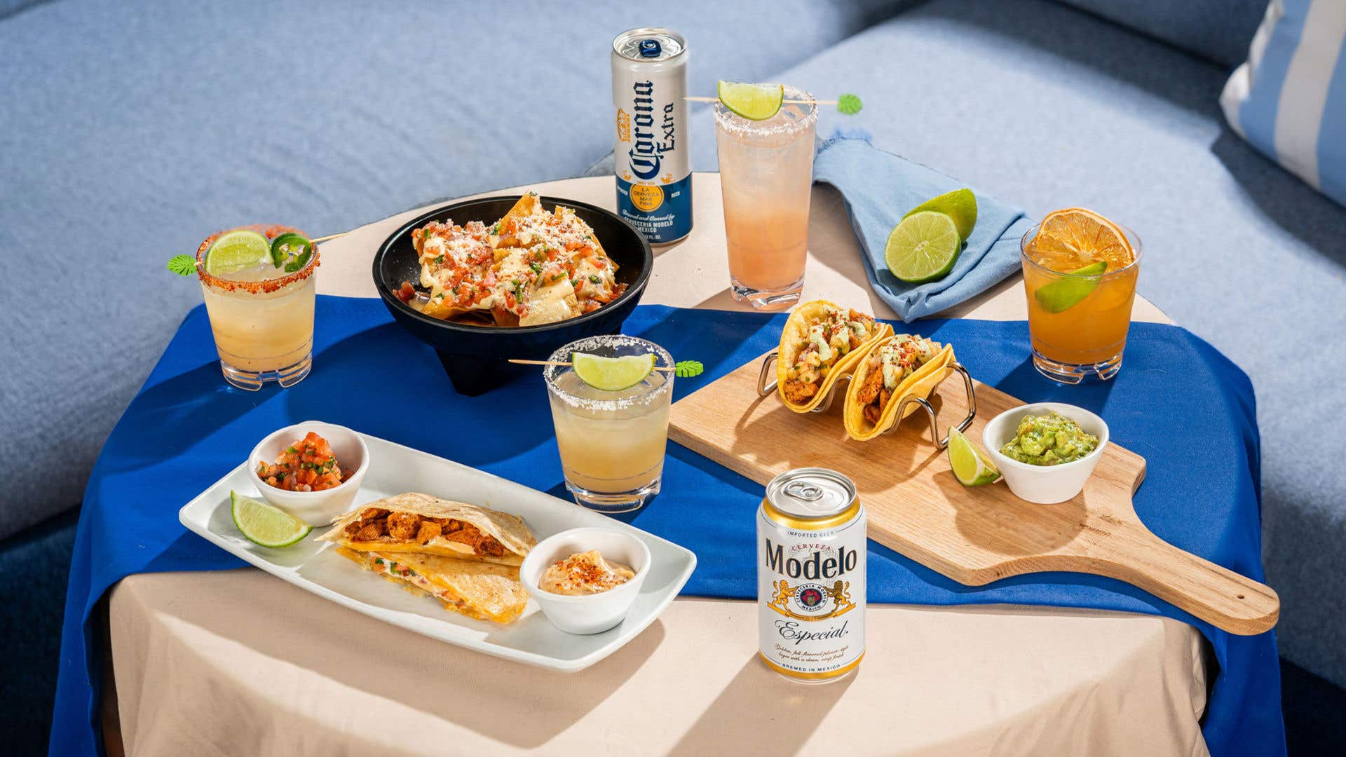 Tabletop spread of food and drinks on a blue and beige table setting. The scene includes tacos, quesadillas, chips and dips, and a bowl of salad, alongside cocktails and cans of Corona Extra and Modelo Especial beer. The items are arranged on platters and cutting boards with lime wedges and small bowls of salsa.