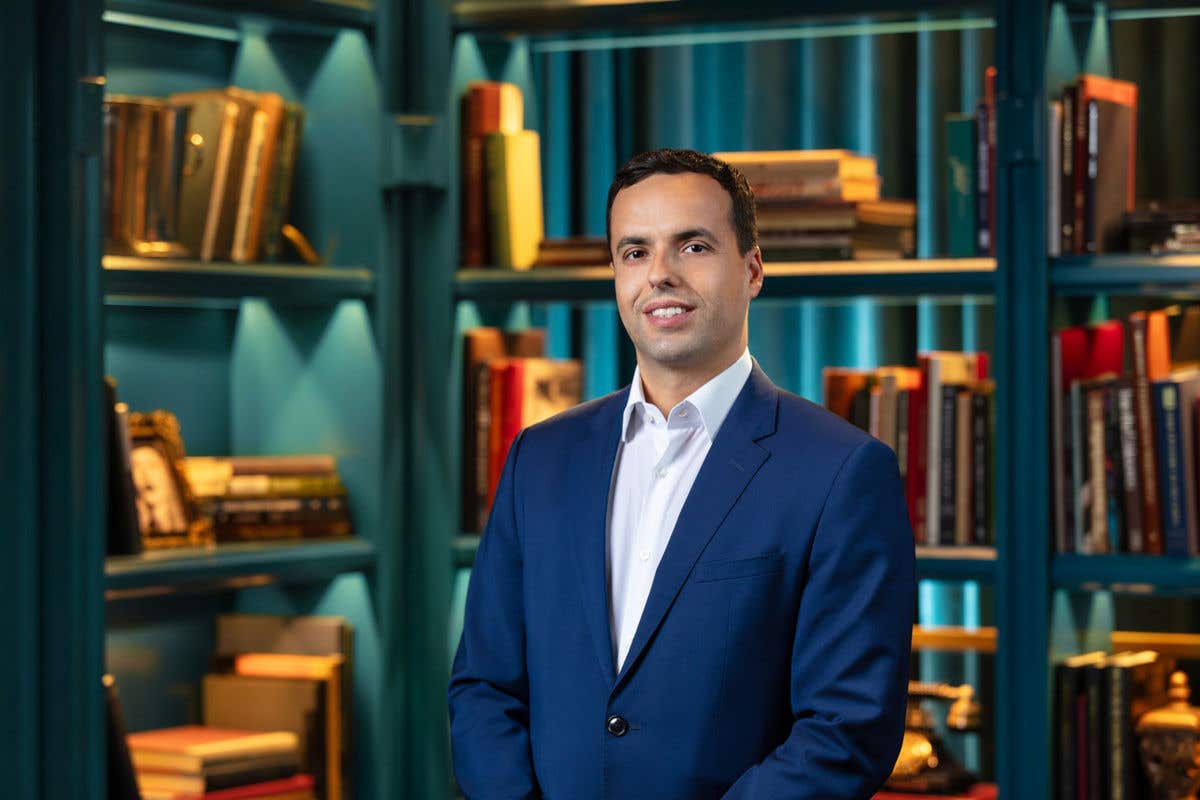 A man with short dark hair, smiling, wearing a blue suit and white shirt, stands in front of a bookshelf.