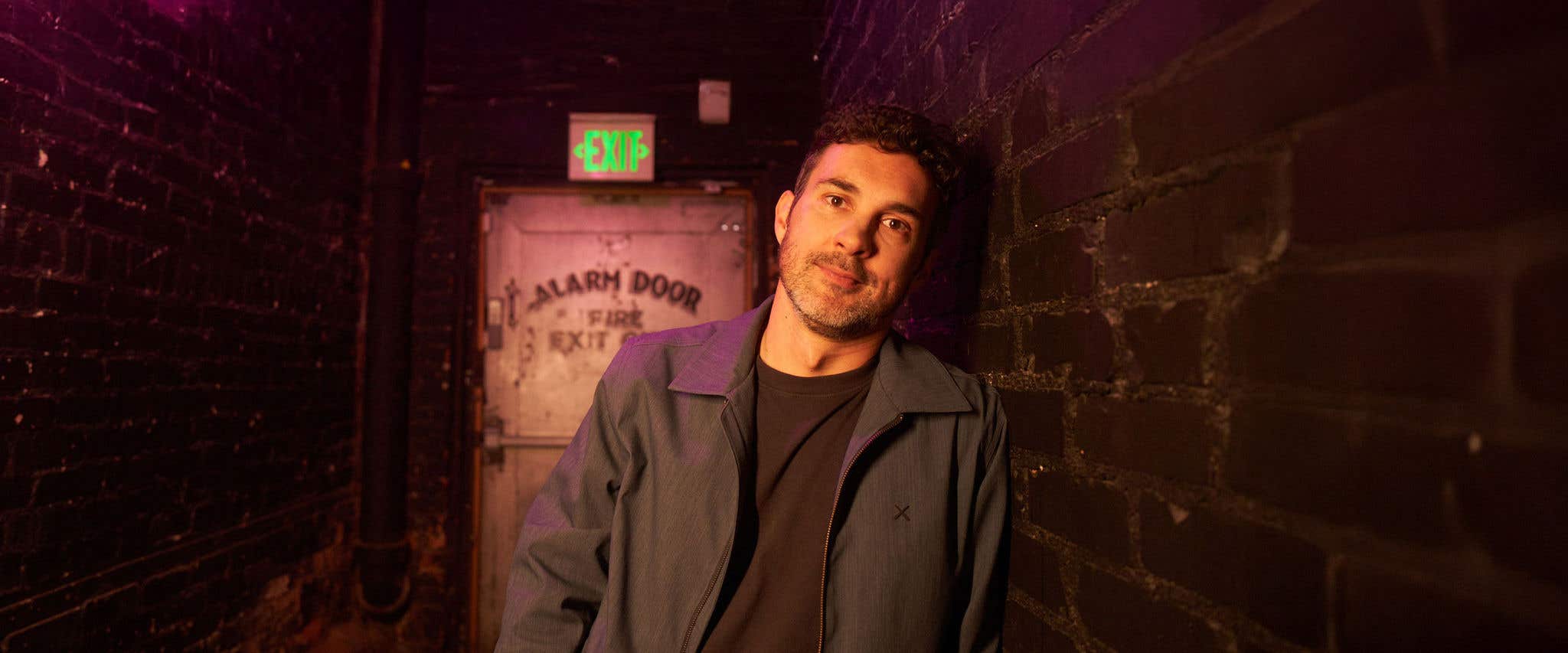 Mark Normand wearing a black shirt and dark jacket leaning against a brick wall looking at the camera. 