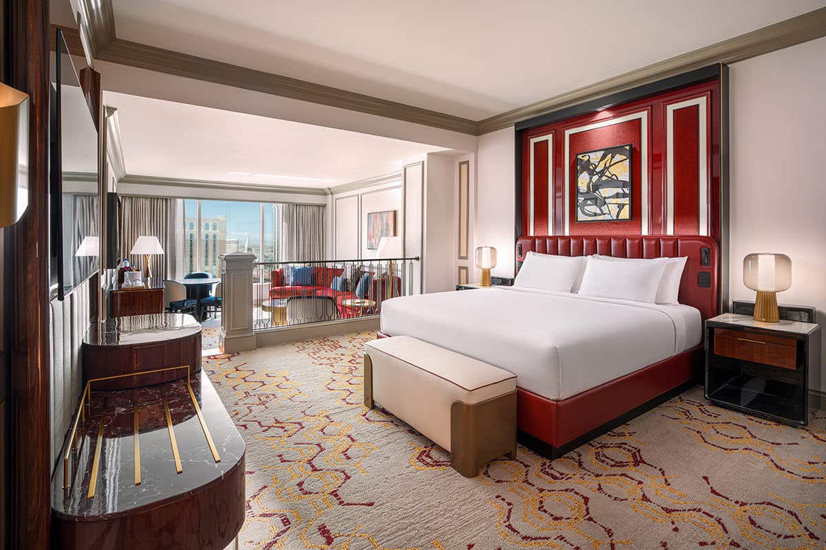 A spacious, split-level hotel suite with a modern and luxurious design. The upper-level bedroom features a king-sized bed with crisp white linens and a striking, tall red leather headboard set against a matching paneled wall. In the foreground, a patterned carpet with red and gold accents covers the floor. A few steps down, a sunken living area is visible past a railing, furnished with a red sofa and offering a bright city view through large windows.