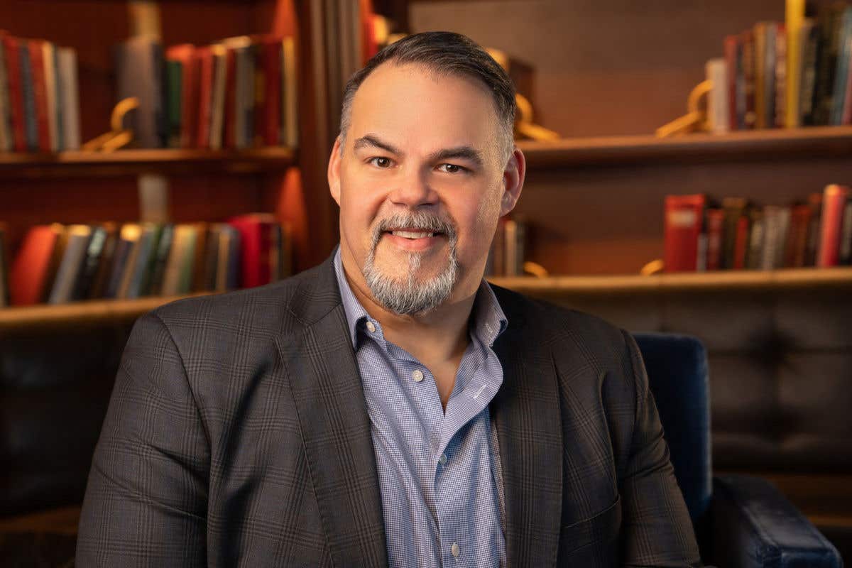 A man with a goatee and short, dark hair, is smiling. He is wearing a blue patterned shirt and a dark blazer and is sitting in a room with bookshelves in the background.
