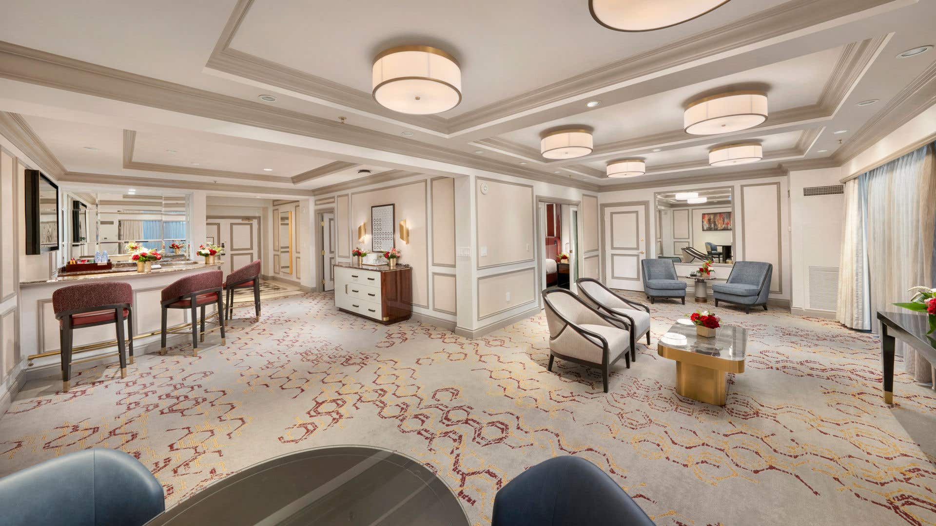 Spacious newly remodeled luxury hotel suite with elegant neutral decor, featuring a large living area, upholstered seating, marble-top coffee tables, a bar with high stools, and soft ambient ceiling lighting.