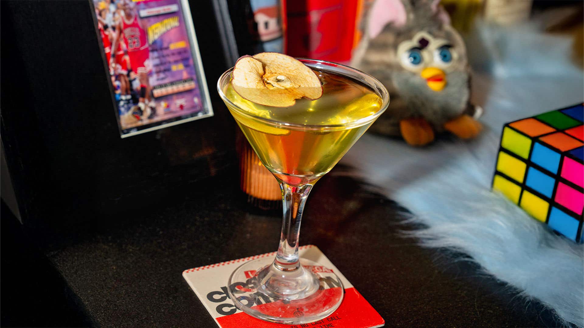 A small martini glass with a dehydrated apple on top. A rubik’s cube and furby are in the background. 