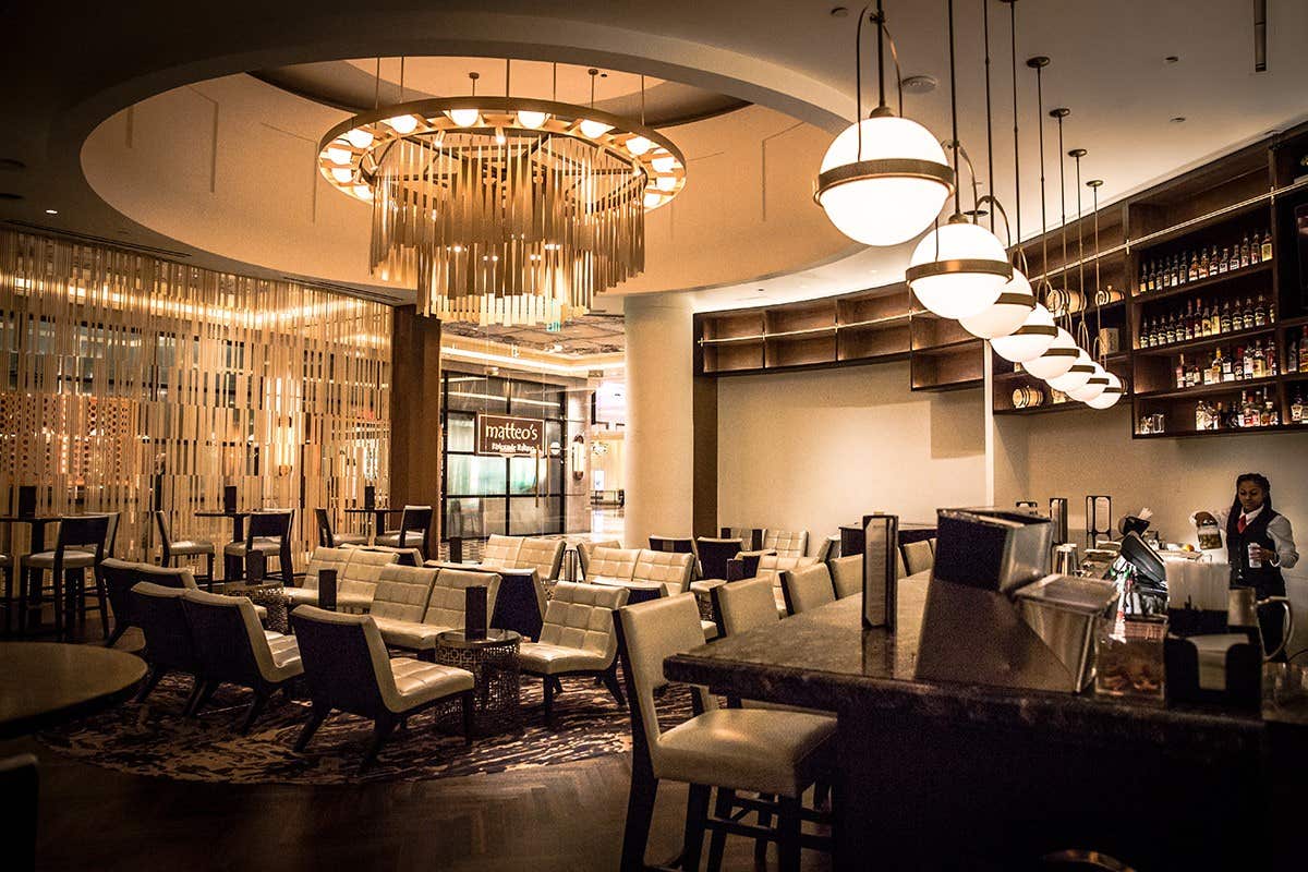 Modern hotel lounge with elegant seating, a large chandelier, round ceiling lights, and a bartender near the liquor shelves.