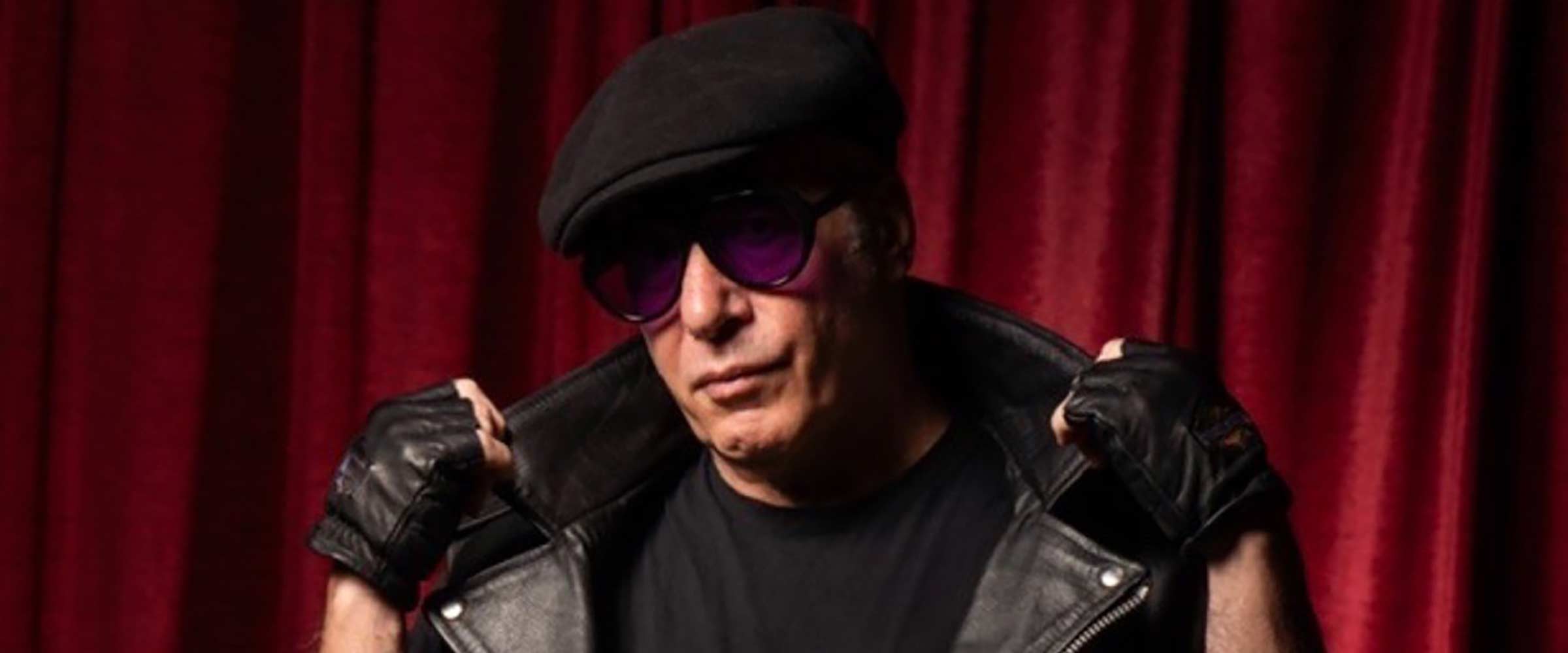Andrew Dice Clay in a black t-shirt and black leather vest wearing leather fingerless gloves, a black beret, dark glasses standing in front of red curtains.  