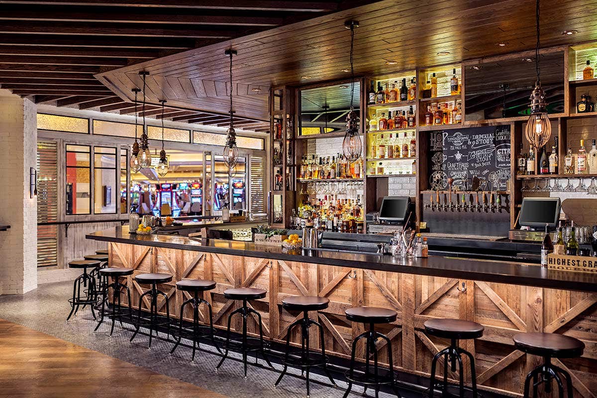 A rustic bar with wooden stools, hanging lights, and a variety of liquor bottles on shelves behind the counter.
