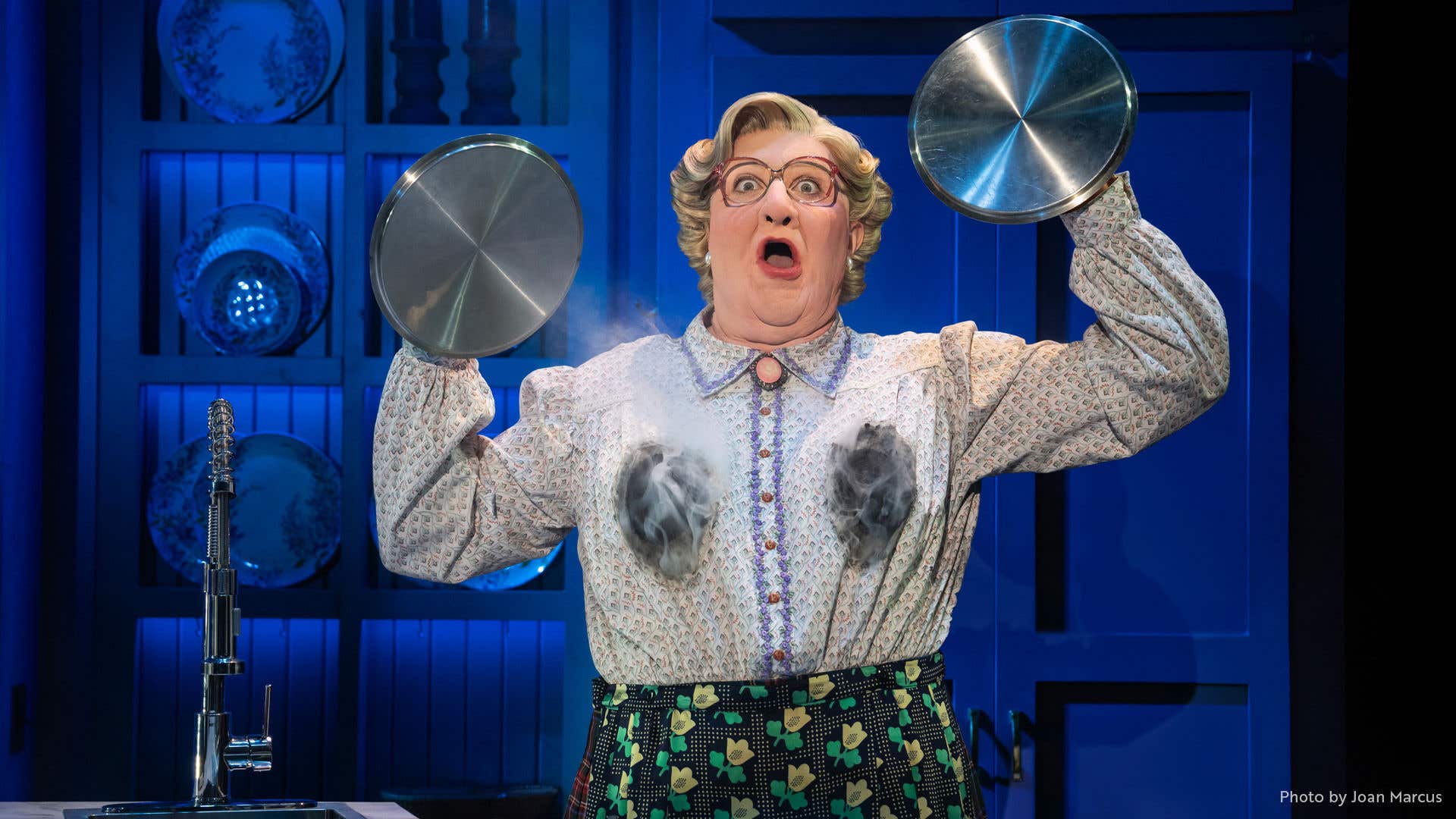 Mrs. Doubtfire holding a pan lid in each hand with a shocked expression. 