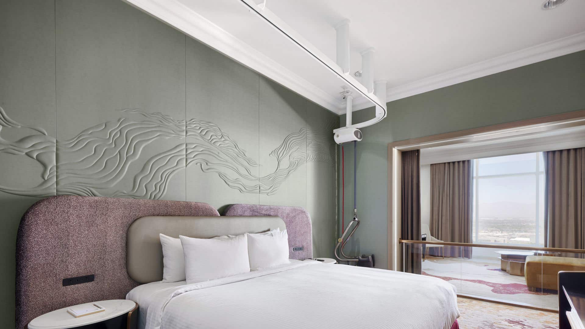 A hotel bedroom with a king bed, padded headboard, abstract wall panels, bedside tables, a mobility lift track in the ceiling going from the corner across the bed, and a glass railing separating the bedroom from the living room with a city view.