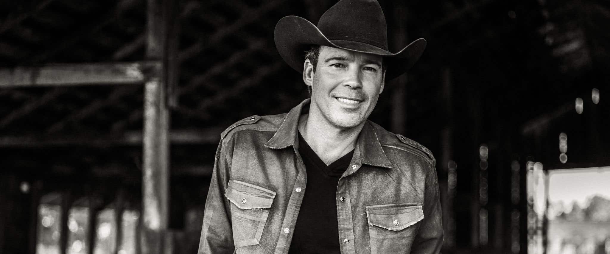 Grayscale image of Clay Walker in a cowboy hat, open button up, black shirt and jeans smiling at the camera. 