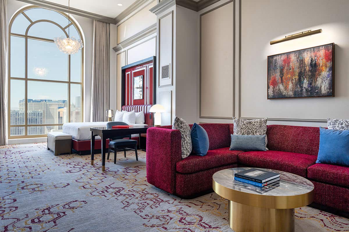A spacious and elegant hotel suite with an open floor plan. In the foreground, a plush, deep-red sectional sofa with decorative pillows sits next to a round, gold coffee table. Beyond the living area, a bedroom space features a neatly made bed with a red leather headboard and a small desk. A large, arched floor-to-ceiling window offers a view of a city skyline during the day. The room is decorated with modern art and features a patterned carpet, creating a luxurious and comfortable atmosphere.