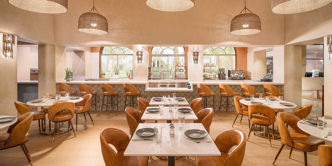 Bright, modern restaurant dining room with rows of square tables set with plates and glassware, surrounded by cushioned tan chairs. Large woven pendant lights hang from the ceiling above, and a bar with high stools lines the back wall beneath arched windows displaying bottles and decor.