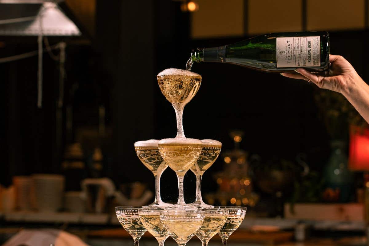 Champagne is poured from a bottle into a pyramid of ornate coupe glasses, creating cascading foam. The glass tower sits in a dimly lit restaurant setting with warm ambient lighting and blurred décor in the background.