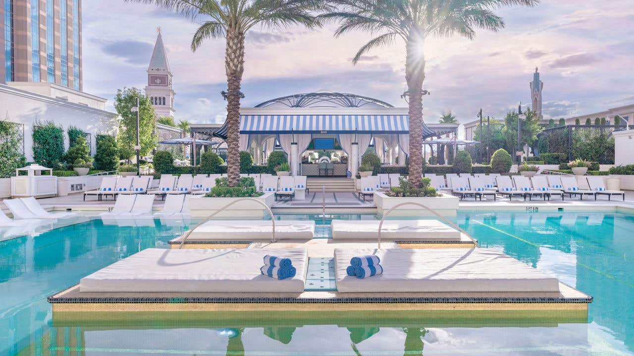 Outdoor hotel pool with two in water loungers, rolled blue and white towels, palm trees, and striped cabanas arranged symmetrically behind the pool.