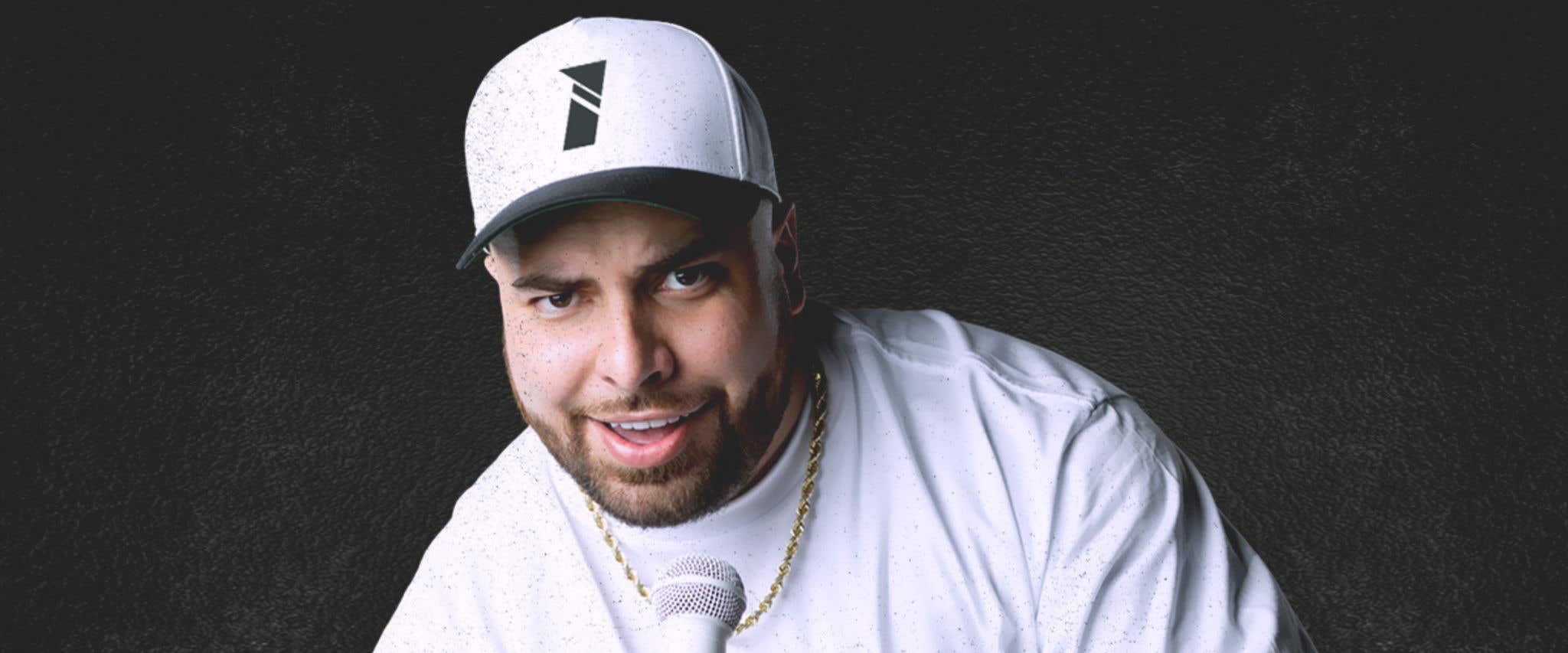Tumua in a white hat, white shirt, wearing a gold chain, and holding a white microphone.