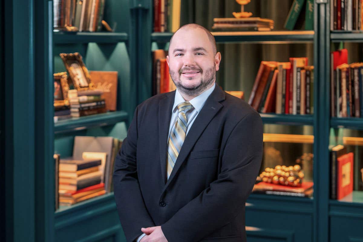 A man with a beard is smiling. He is wearing a dark suit, a light-colored shirt, and a diagonally striped tie. He is standing in front of a blue-green bookshelf filled with books. 