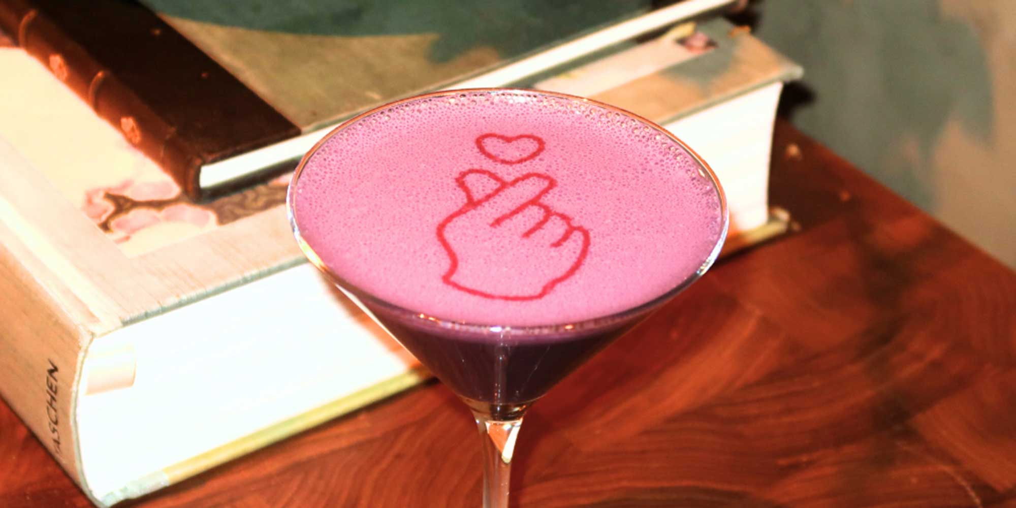A purple cocktail in a martini glass, topped with pink foam that has a red finger heart design stenciled on it.