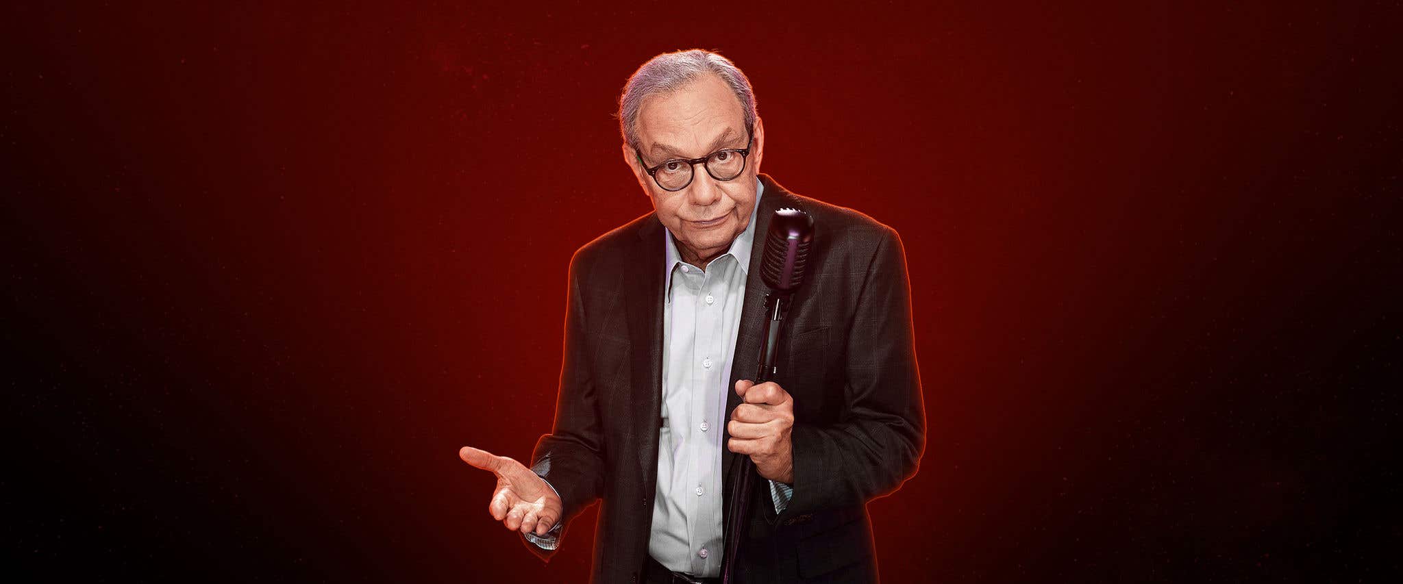 Lewis Black holding a mic stand in front of a dark red background looking at the camera. 