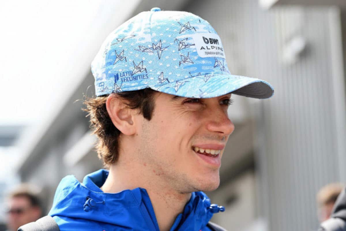 Close-up of a person wearing a light blue Alpine team cap and blue jacket, photographed outdoors with buildings blurred in the background.