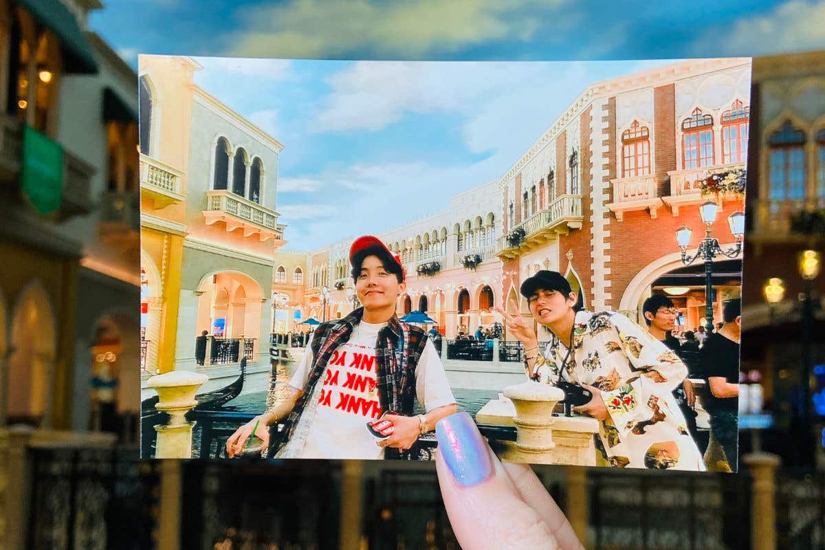 A hand holding a photograph of two men posing in front of the Grand Canal Shoppes, aligned with the real-life background.