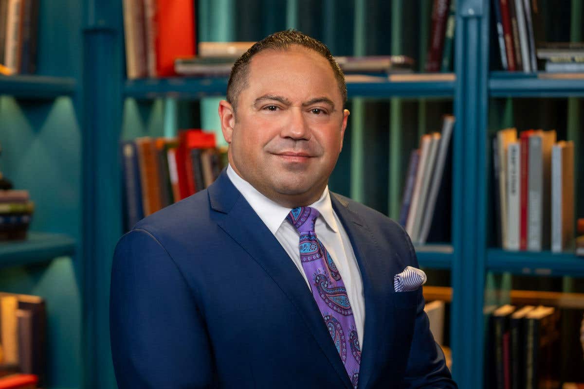 A man with dark, slicked-back hair is smiling. He is wearing a blue suit, white shirt, and a purple paisley tie, and is standing in front of a blue-green bookshelf.