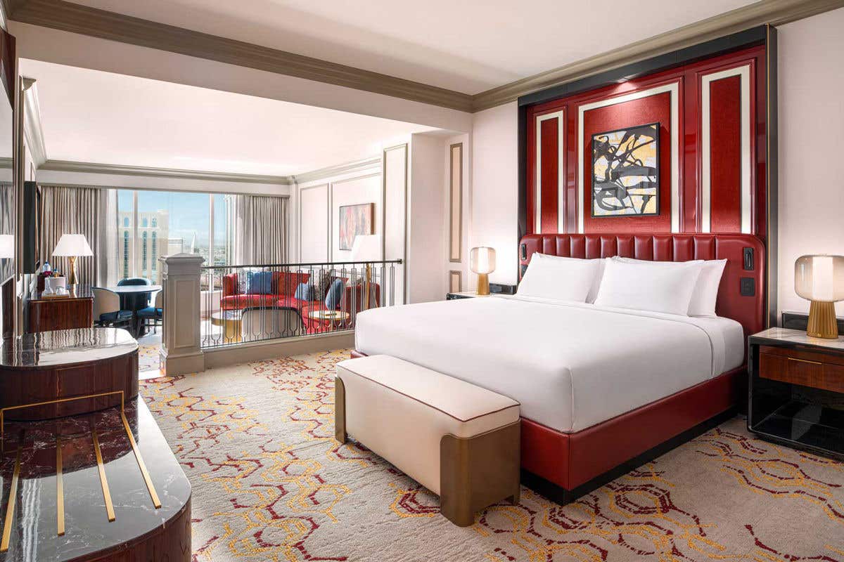 Luxurious hotel room with a king-sized bed, red headboard, abstract artwork, red sectional, and large window.
