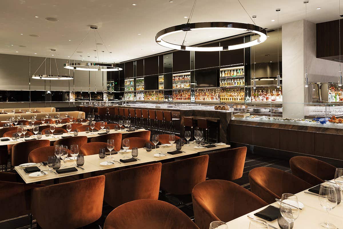 Modern restaurant interior with elegant lighting, a well-stocked bar, and neatly arranged tables with brown chairs.