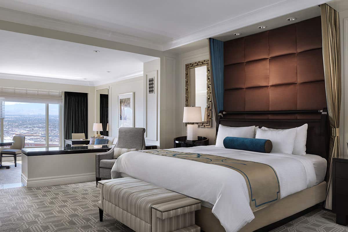 A luxurious hotel suite with a large, comfortable king-sized bed as the centerpiece. The bed features a tall, tufted brown headboard, crisp white linens, and a decorative runner. To the left, a sunken living area with a desk and chairs offers a panoramic view of a city and mountains through a large window. The room's elegant design, combining modern furniture and a soft, neutral color palette, creates a serene and upscale atmosphere.