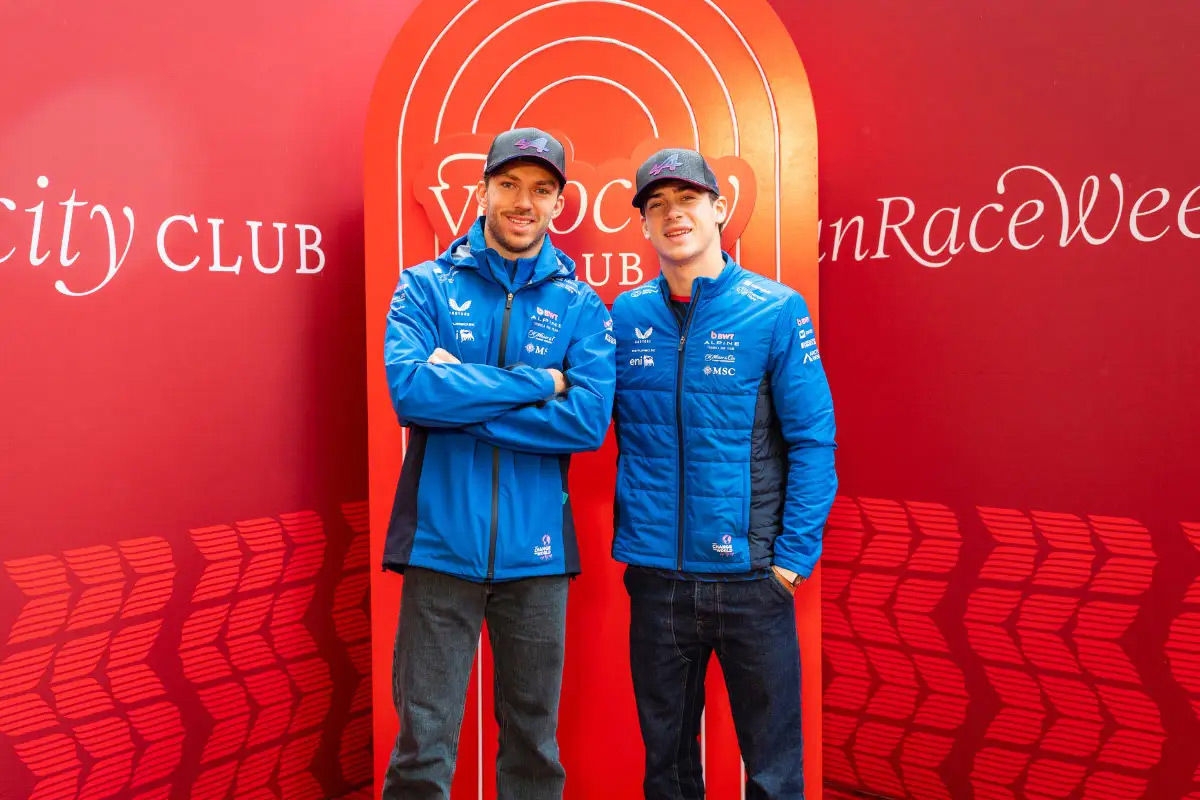 Two race drivers wearing blue team jackets and caps stand in front of a red backdrop.