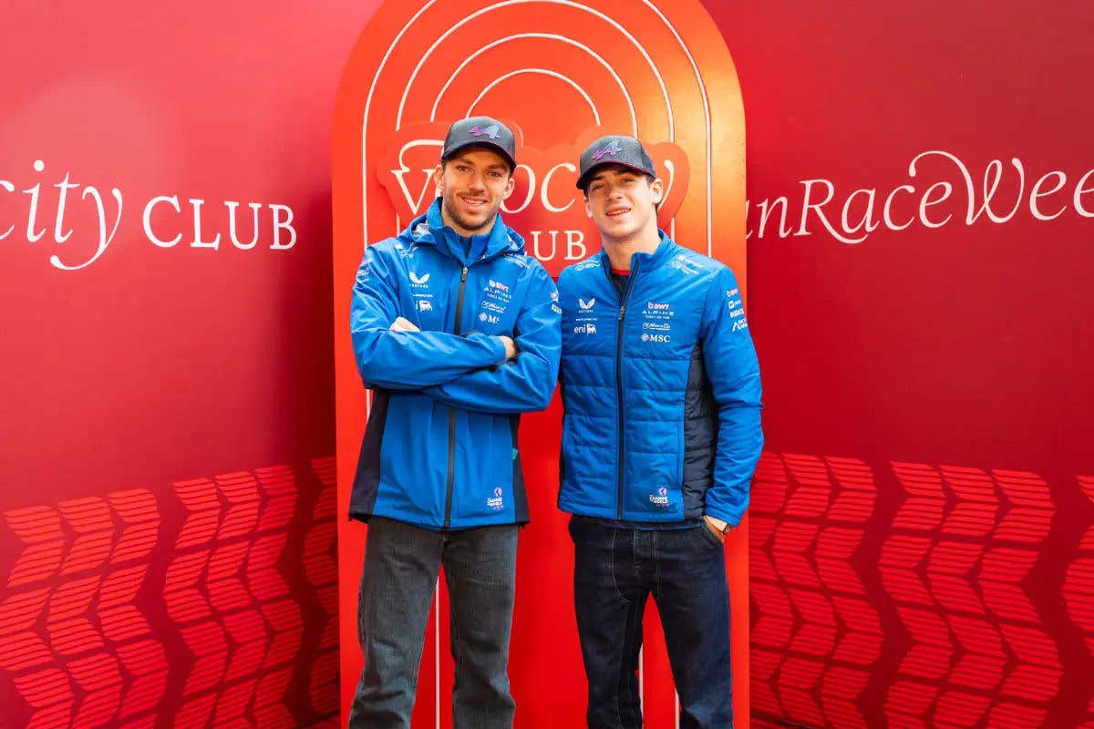 Two race drivers wearing blue team jackets and caps stand in front of a red backdrop.