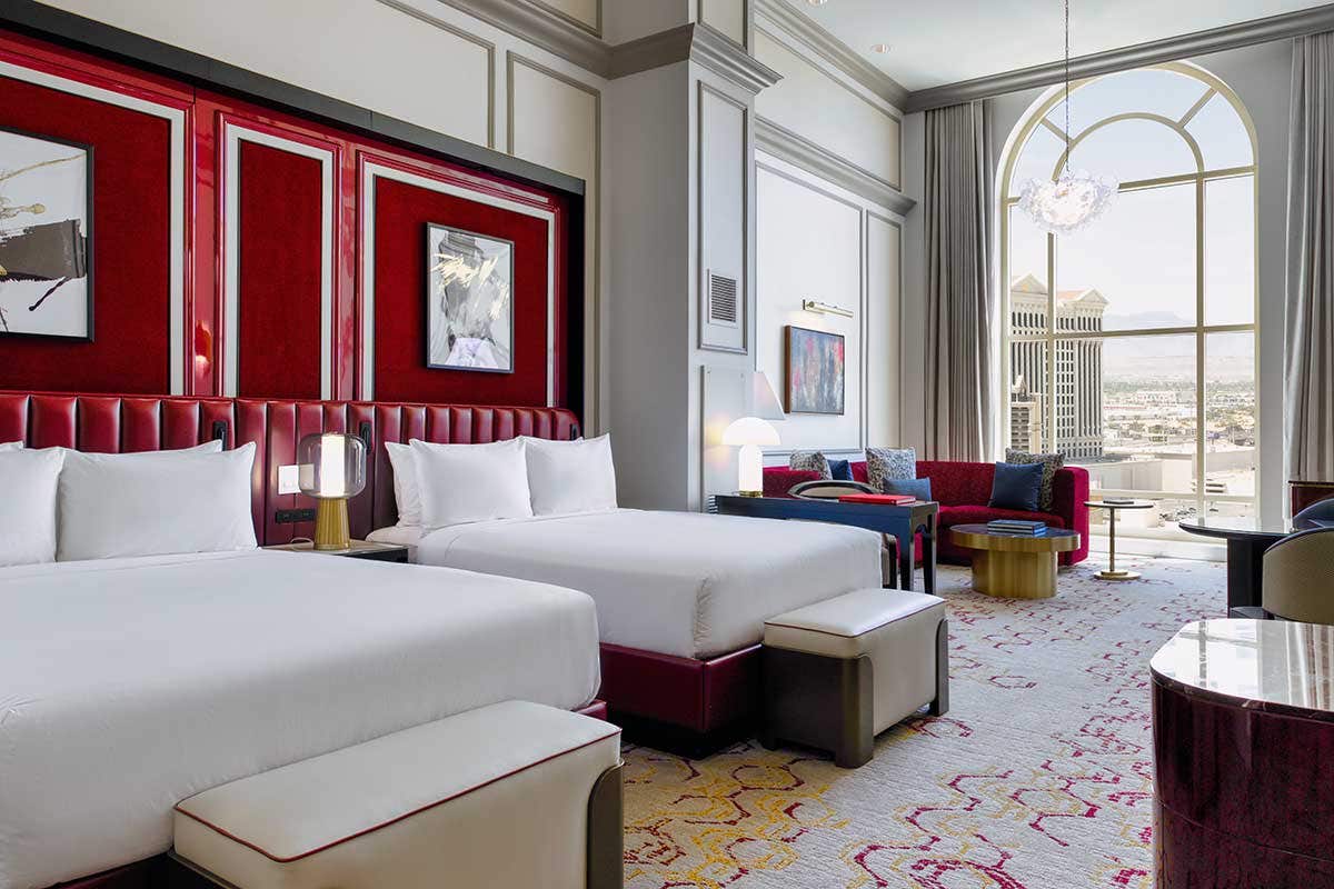 A hotel room with two queen beds, an armchair, a red curved couch, a gold coffee table, and a large arched window.