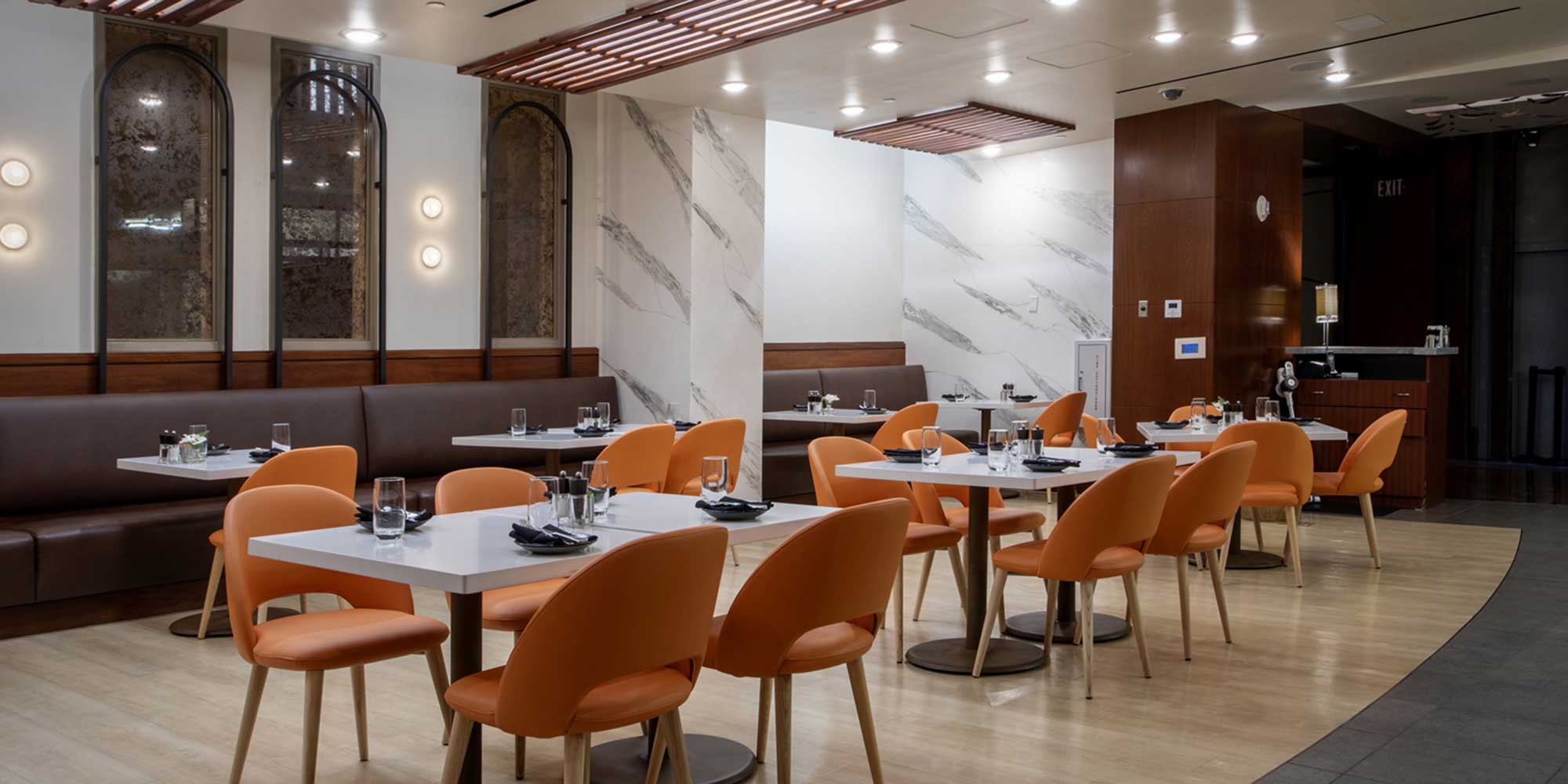 Modern restaurant dining area with white tables, orange chairs, a brown banquette, and warm lighting on marble-accented walls.