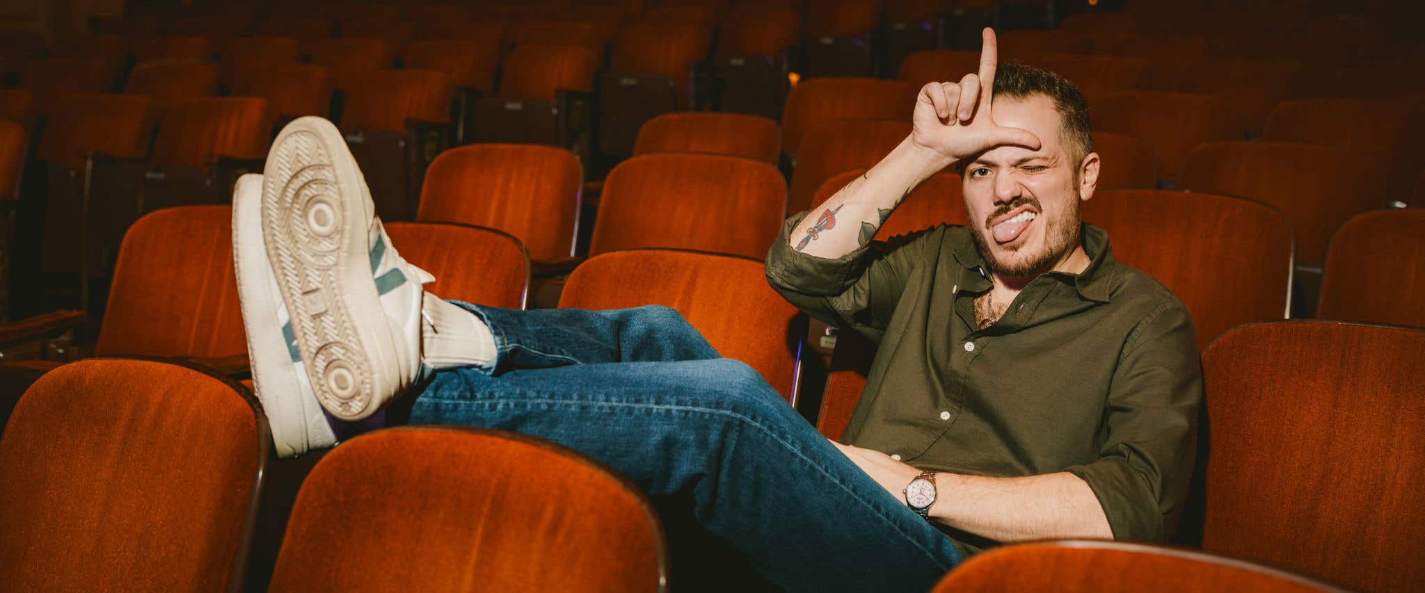 Jeff Arcuri sitting in theater seats with his feet propped up on the seat in front of him looking at the camera with his tongue out and his hand making an L shape on his forehead.  