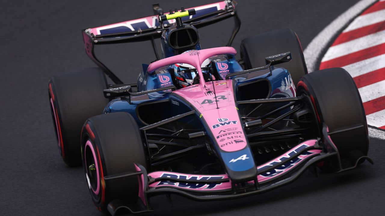 Close-up front view of a pink and blue Formula 1 race car on a racetrack, showing the front wing, tires, and driver’s helmet inside the cockpit.