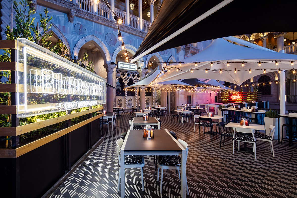 An outdoor restaurant with string lights, tiled flooring, and white tables under large umbrellas in a European-style setting.