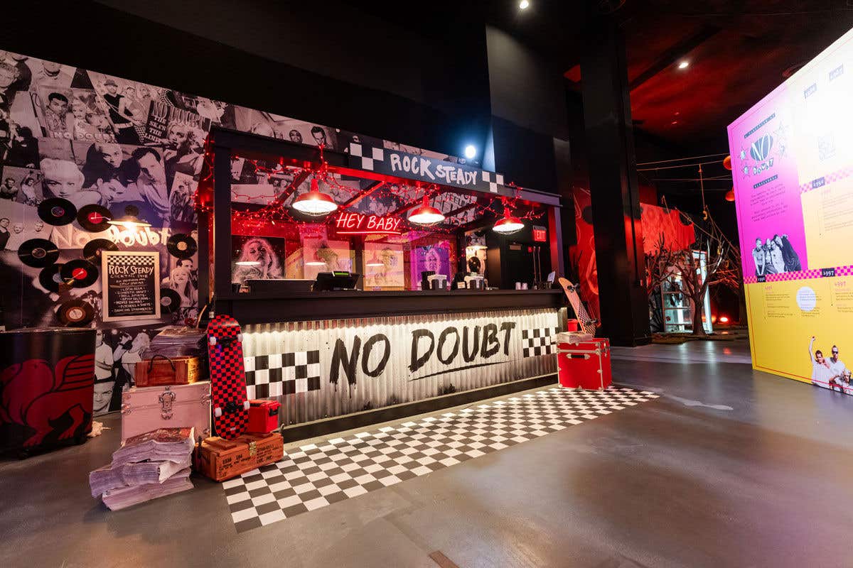 A black and white checkered concert merchandise stand that says “Rock Steady,” “No Doubt” and “Hey Baby” with black and white photos behind and red music cases.