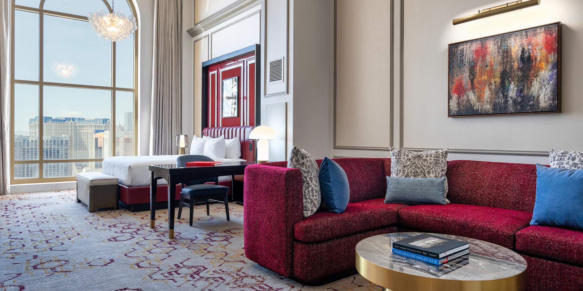 A spacious and elegant hotel suite with an open floor plan. In the foreground, a plush, deep-red sectional sofa with decorative pillows sits next to a round, gold coffee table. Beyond the living area, a bedroom space features a neatly made bed with a red leather headboard and a small desk. A large, arched floor-to-ceiling window offers a view of a city skyline during the day. The room is decorated with modern art and features a patterned carpet, creating a luxurious and comfortable atmosphere.