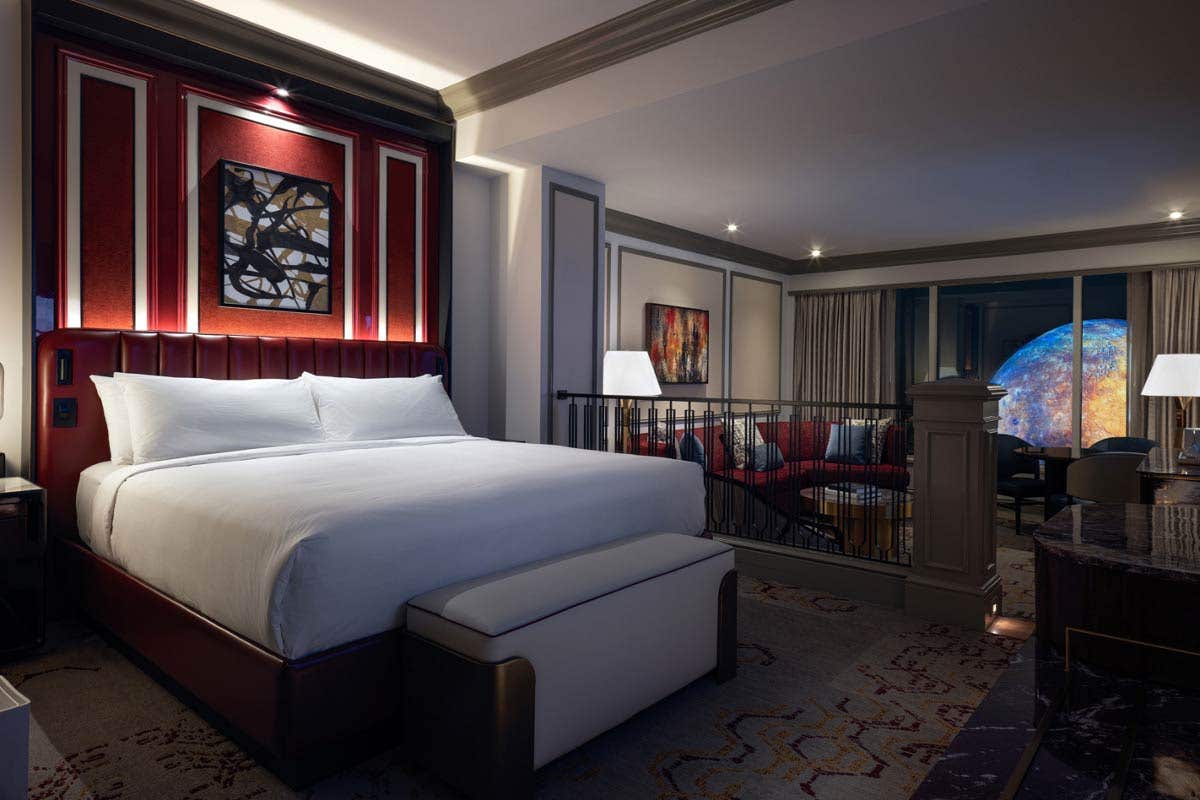 Luxury King Suite bedroom with a bench seat at the foot of the bed. In the background is a sunken living room with an armchair, couch, coffee table, dining table with chairs, lamps, and a nighttime view of Sphere at The Venetian outside the window.