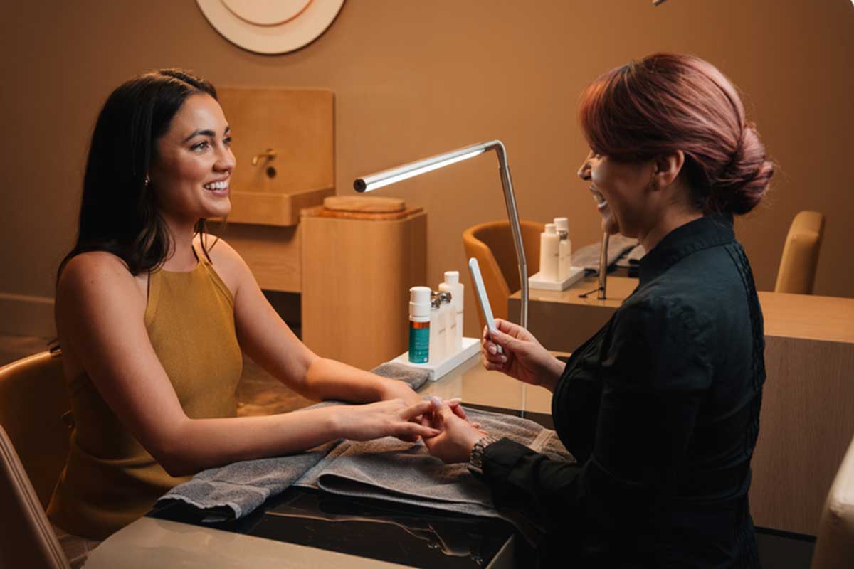 Two people seated across a manicure table in a softly lit spa setting, with one person’s hands resting on a towel while the other holds a manicure tool. A desk lamp, nail care bottles, and small containers are arranged on the table, with neutral-toned walls and cabinetry in the background.
