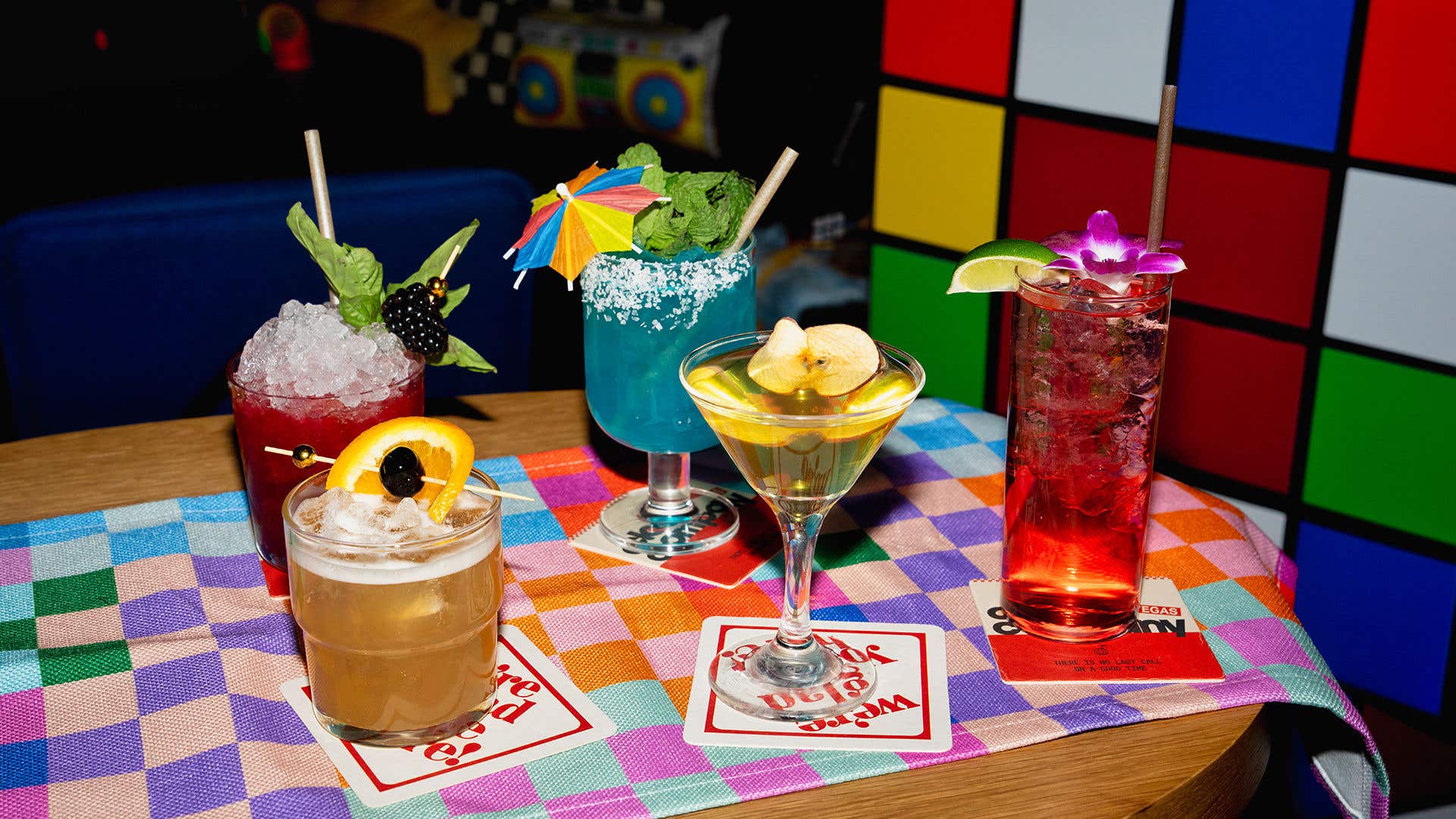 Five brightly colored cocktails in a variety of glassware on a table. An oversized rubik’s cube and boombox are visible in the background.