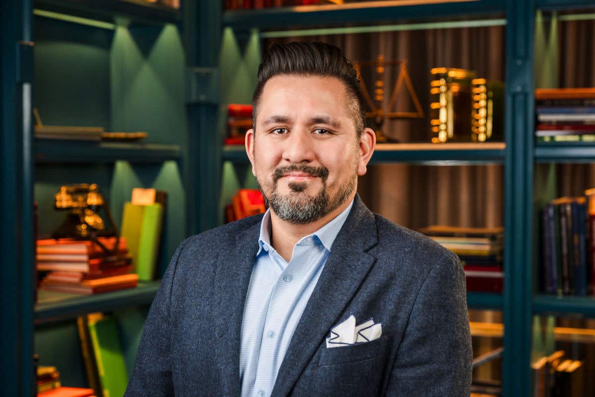 A man with dark hair styled up, a short beard, and a mustache is smiling. He is wearing a light blue collared shirt and a dark tweed blazer with a white pocket square and is in a room with blue-green bookshelves in the background.