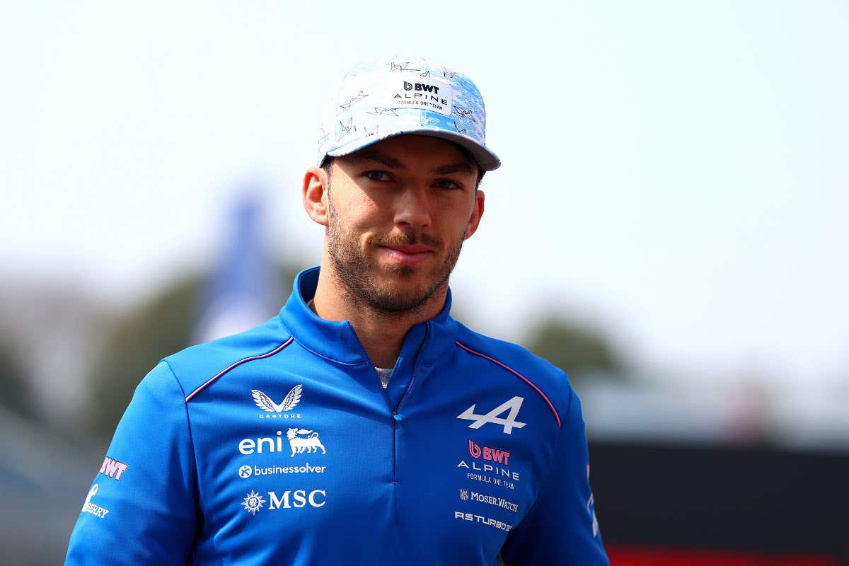 Medium close-up of a person wearing a blue Alpine team jacket with sponsor logos and a matching light blue cap, standing outdoors with a racetrack-style background out of focus.