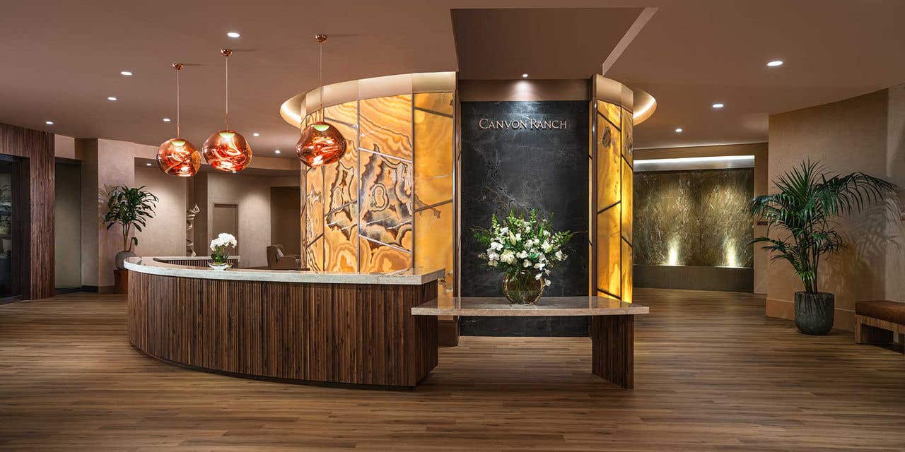 Spa reception area with a curved wood front desk, amber stone and black marble wall panels labeled “Canyon Ranch,” warm pendant lighting, wood flooring, and potted plants in a modern, softly lit interior.