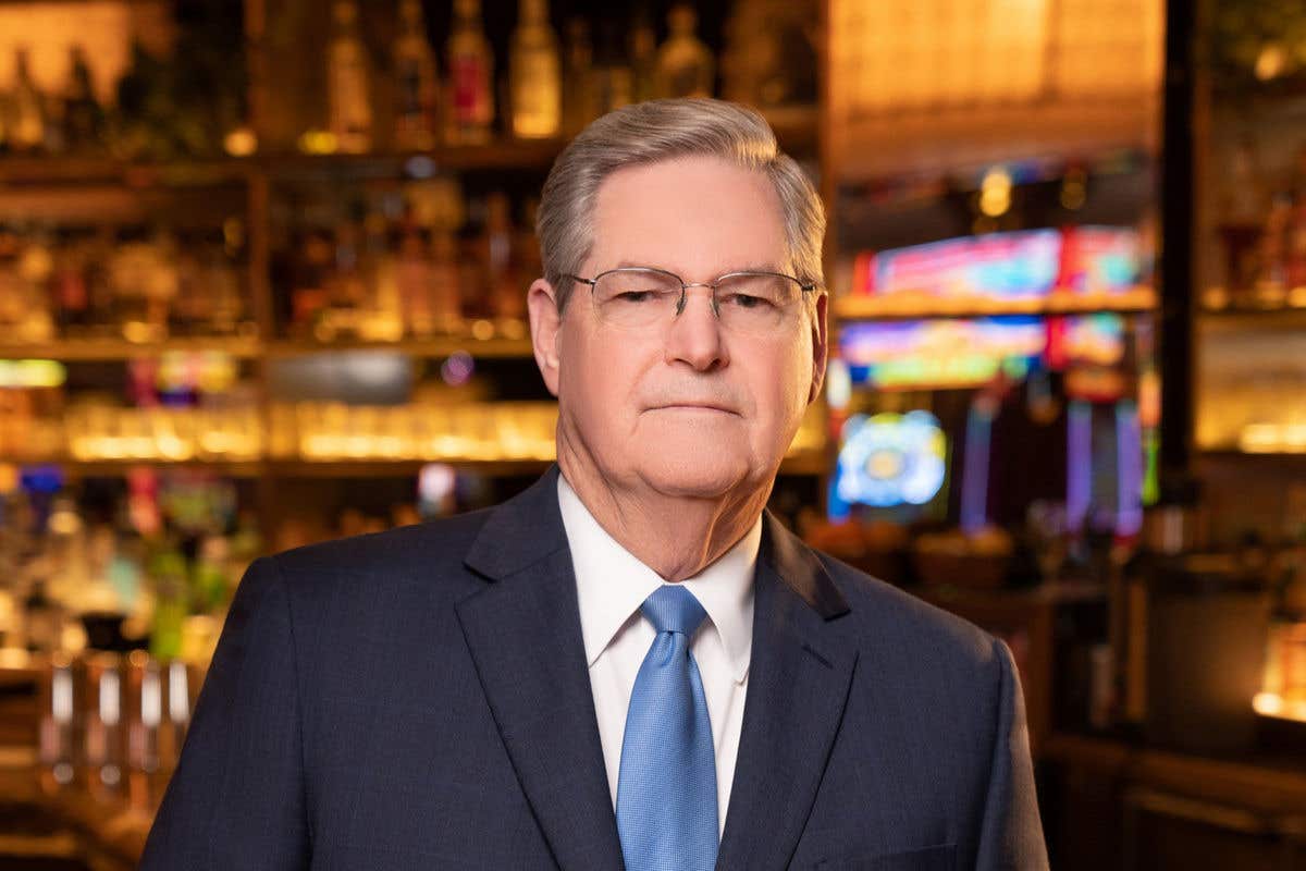 A man with glasses and graying hair, wearing a dark suit and a blue tie, stands in front of a warmly lit bar.