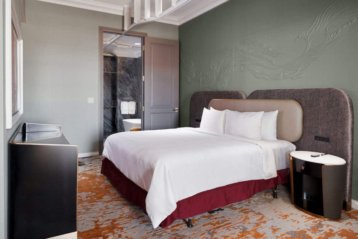 A hotel bedroom with a king bed, padded headboard, side tables, and an open doorway leading to a marble bathroom with a mobility lift track in the ceiling that goes across the bed and into the bathroom.
