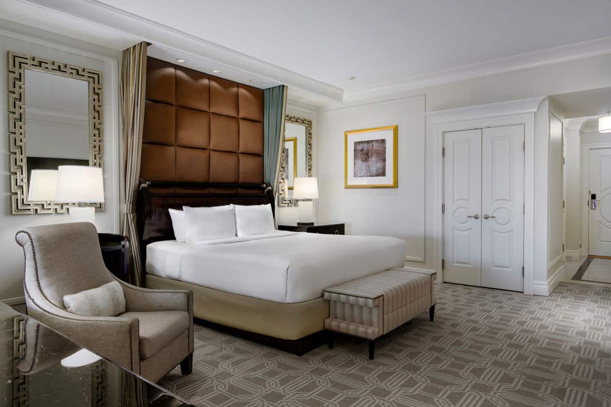 Elegant hotel bedroom featuring a king bed with a tufted leather headboard, upholstered chair, bench, mirrored accents, and soft neutral décor.