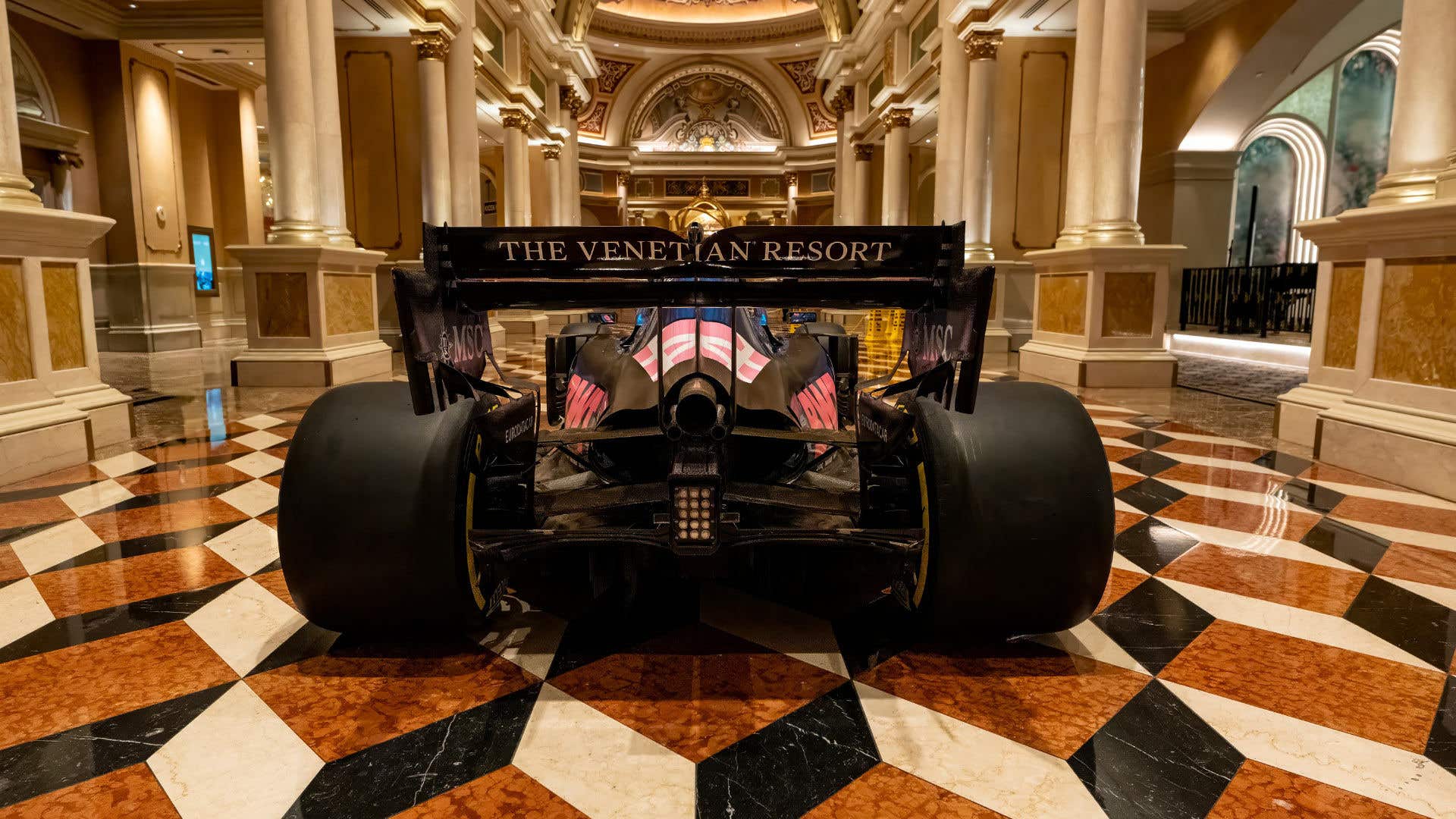 Rear view of a pink and blue Formula 1 race car displayed indoors on a marble floor, with “The Venetian Resort” visible on the rear wing inside a grand column-lined hallway.