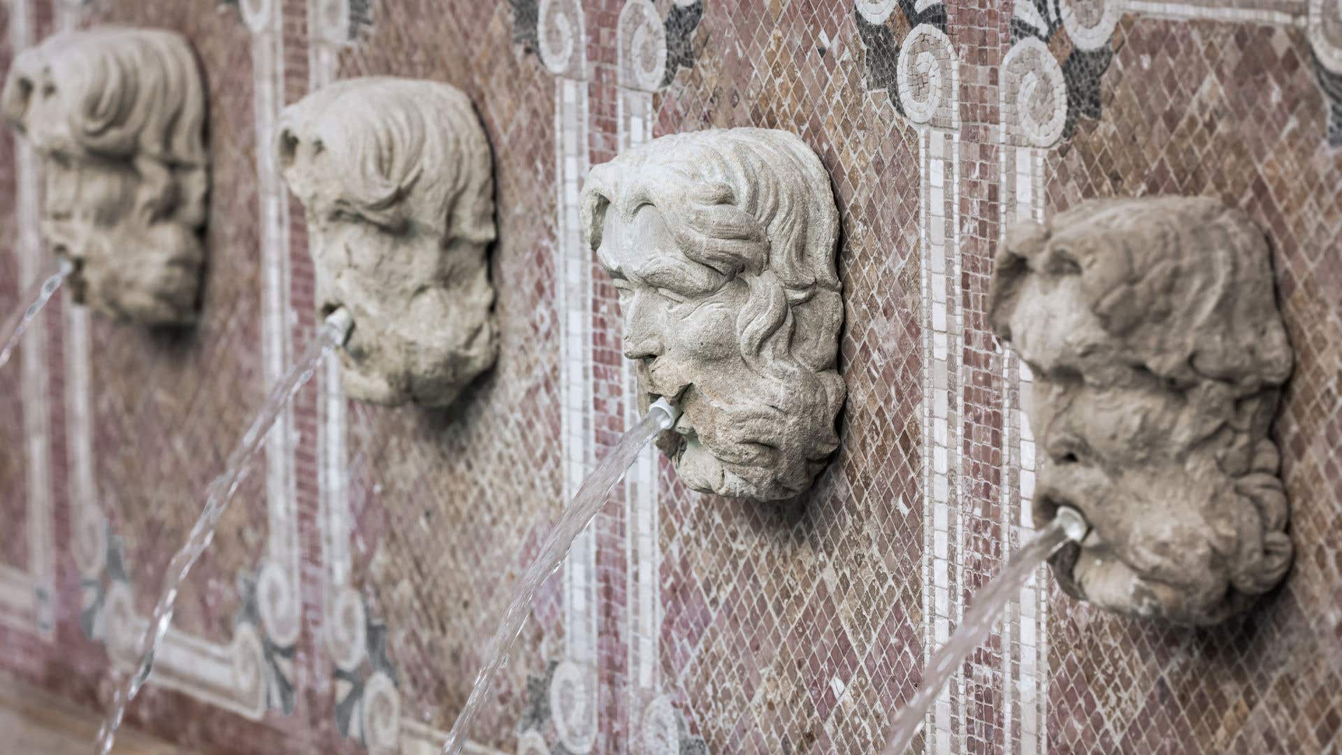 Stone wall fountain featuring four sculpted heads arranged in a row, each with a stream of water flowing from the mouth into a basin below, set against a mosaic tile background.