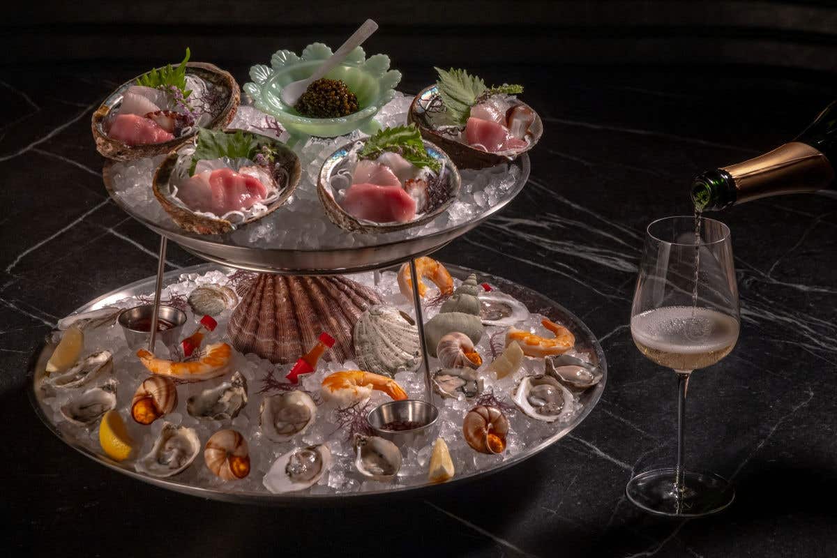 Tiered seafood platter on crushed ice featuring oysters, shrimp, shellfish, and sashimi served in shells, with caviar in a bowl at the center. Sparkling wine is being poured into a glass beside the display on a dark marble table.