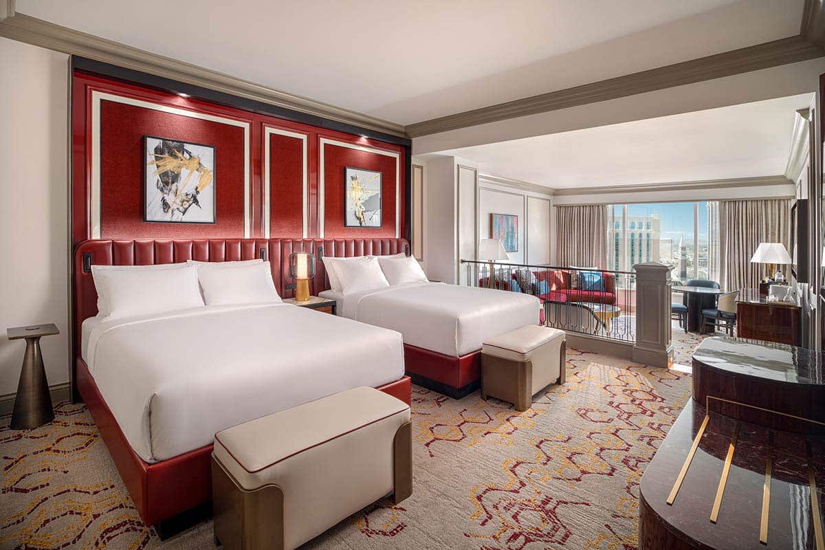 Remodeled Luxury Two Queen Suite bedroom with a bench seat at the foot of the bed. In the background is a sunken living room with an armchair, couch, coffee table, dining table with chairs, lamps, and a daytime view outside the window.